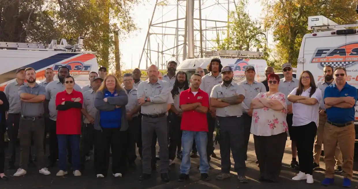 A group of people in work uniforms and casual clothes stand with arms crossed in front of company vehicles and equipment outdoors.