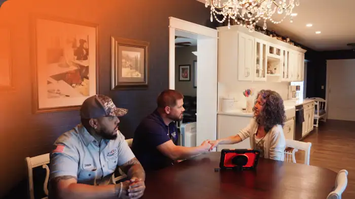 Two men sit at a dining table with a woman in a kitchen; one man and the woman are shaking hands, and a tablet is on the table.