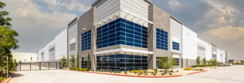 Modern industrial warehouse with large glass windows, multiple stories, and a landscaped entrance, situated under a partly cloudy sky.